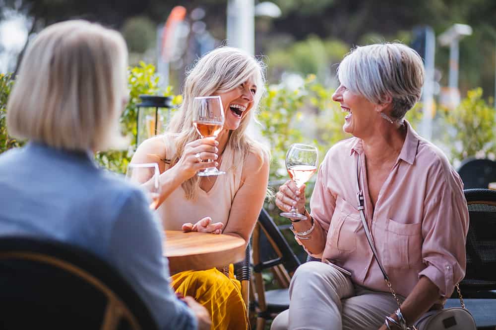 Active adults sitting at an outdoor dining area table. They are laughing while holding glasses of wine.