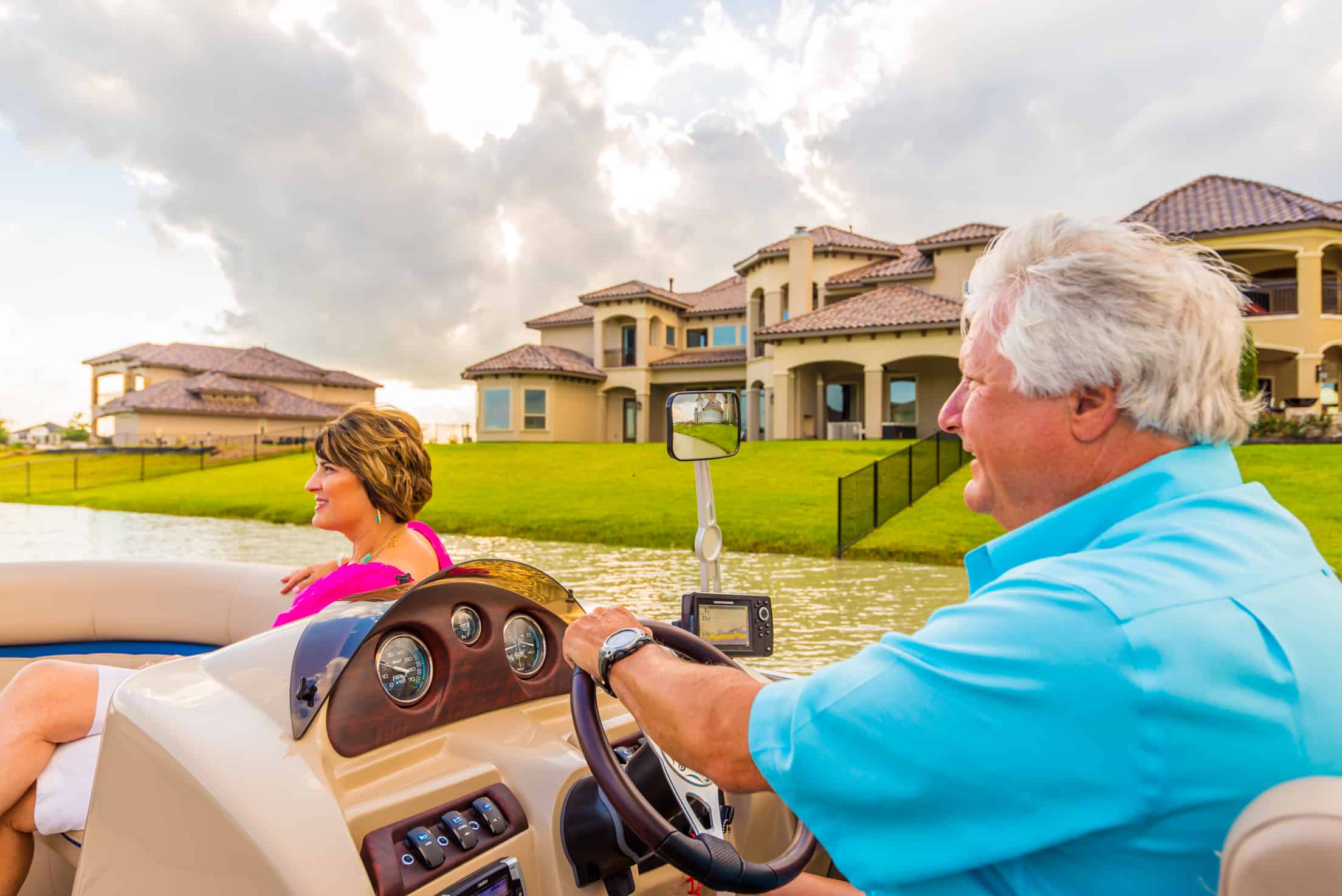 Two people riding a boat near The Cadence Creek at Towne Lake apartments, featuring a view of the lawns and the buildings.